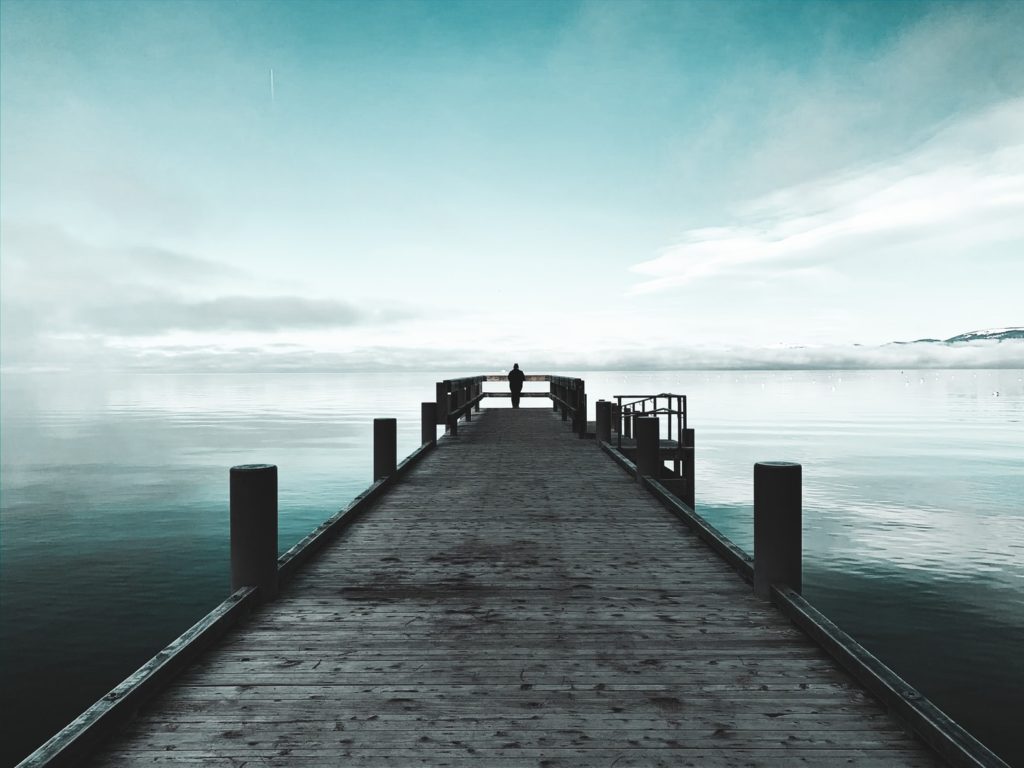 A person stands on the far end of a dock that stretches out into the open sea, symbolizing loneliness.